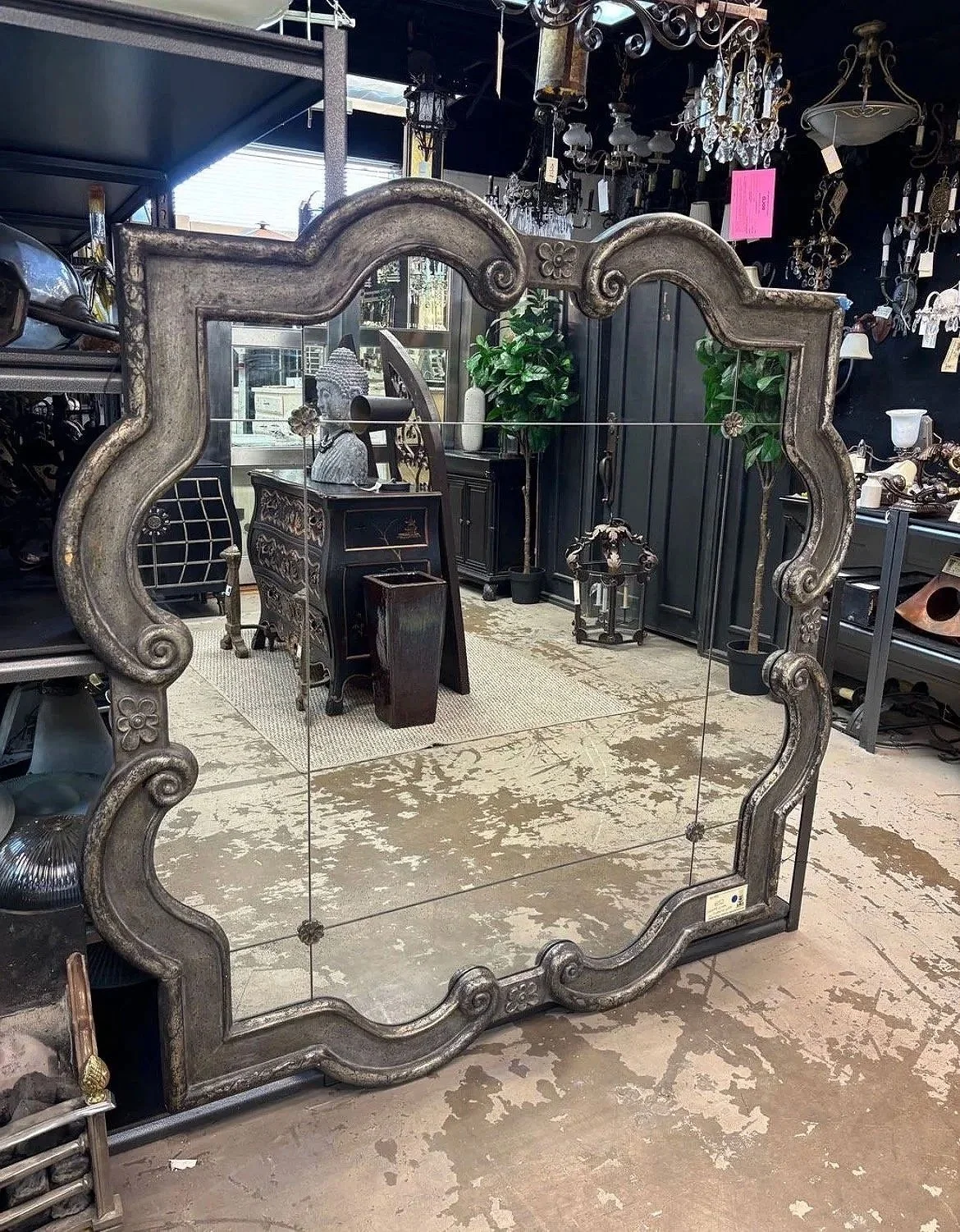 An antique mirror with a decorative frame in a store, reflecting various chandeliers and furniture.