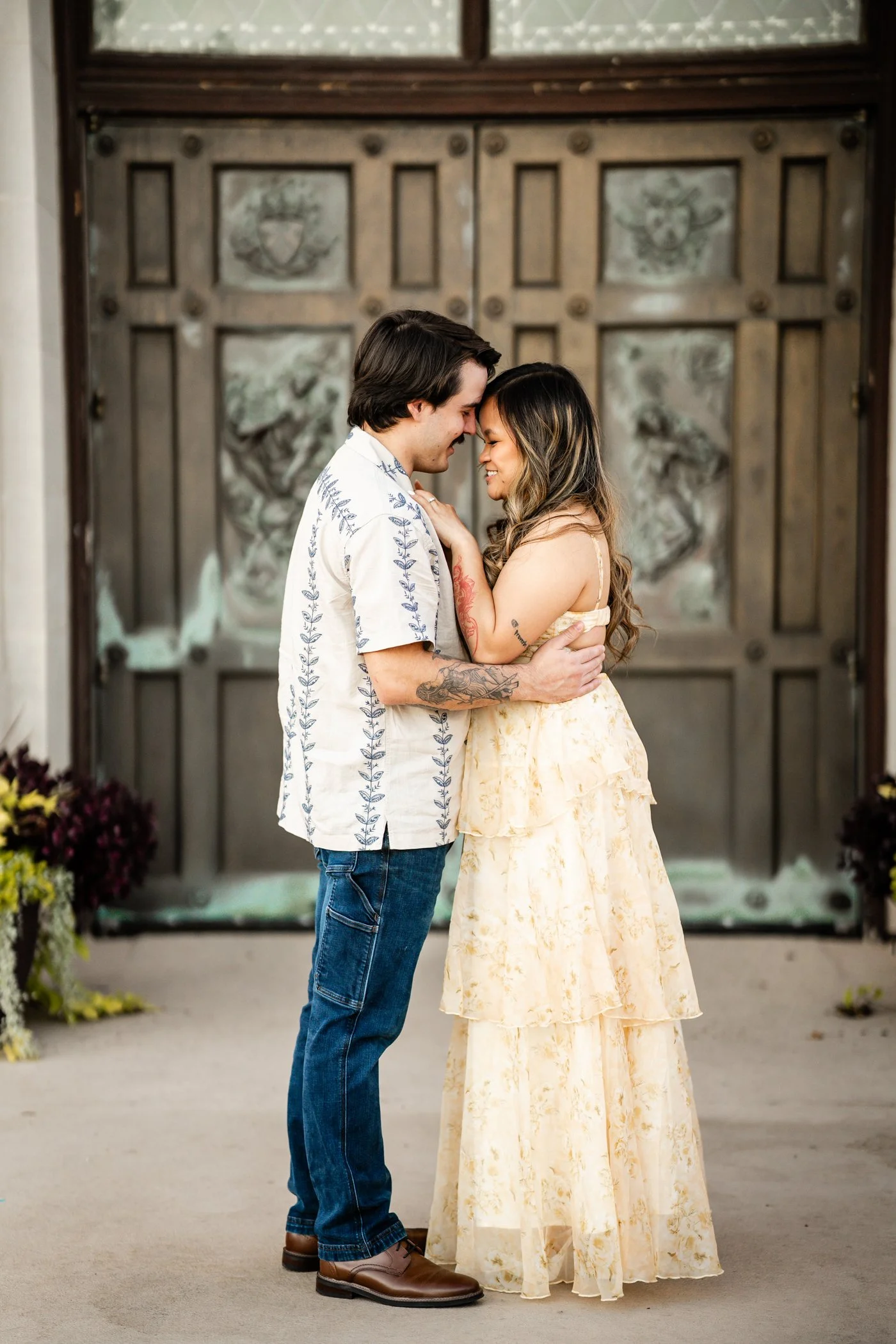 A couple stands close together, touching foreheads and smiling, in front of a large wooden door with decorative panels, surrounded by potted plants. Jevon Photo is based out of Portage, WI and the surrounding areas.