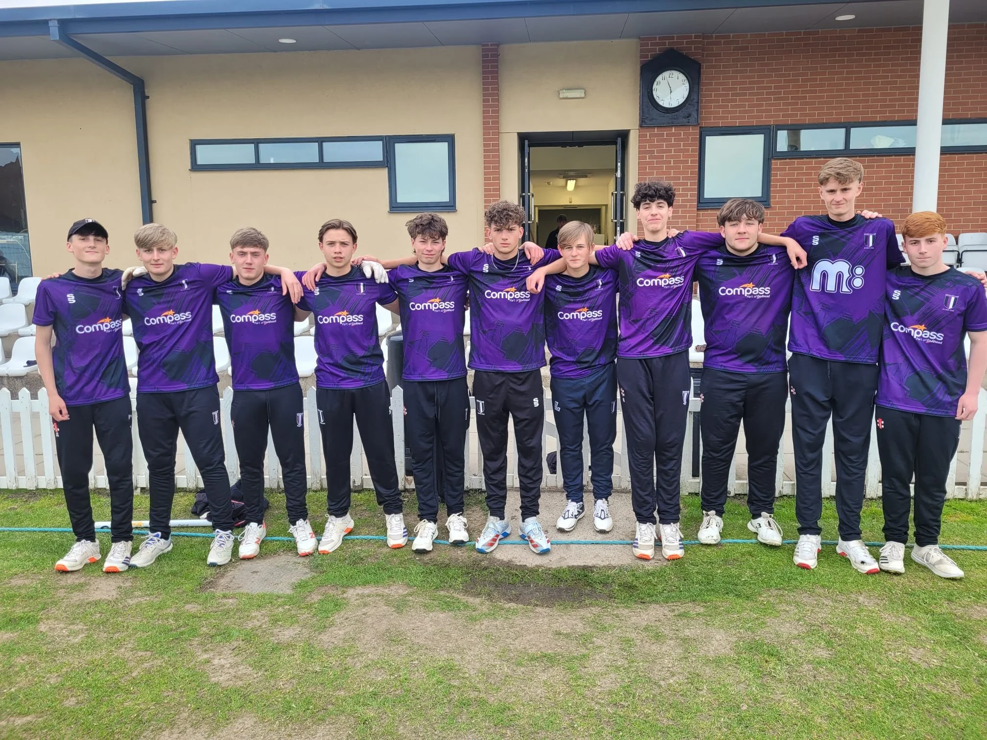 A group of ten young men standing side by side outdoors, with their arms around each other's shoulders, wearing matching purple sports jerseys and black athletic pants, in front of a beige and brick building with a clock showing approximately 3:09.