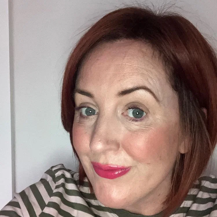 Close-up of a woman with short red hair, blue eyes, and pink lipstick, smiling at the camera in front of a white wall.