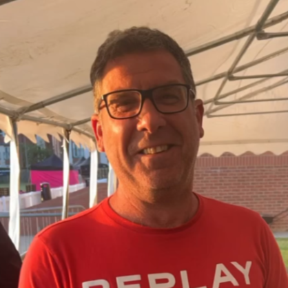 Man with dark hair, glasses, and a red shirt smiling, standing outdoors under a canopy.