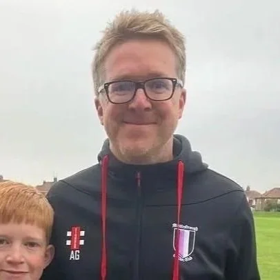A man wearing glasses and a black jacket standing outdoors on a cloudy day, with a partially visible child with red hair next to him.
