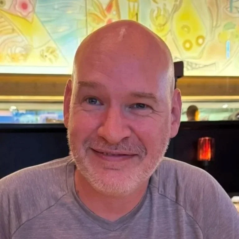 A middle-aged man with a bald head and gray beard smiling at the camera in a restaurant setting, with colorful abstract art on the wall behind him.