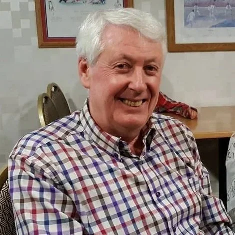 A smiling senior man with white hair wearing a plaid shirt, sitting indoors with a framed picture and chairs in the background.