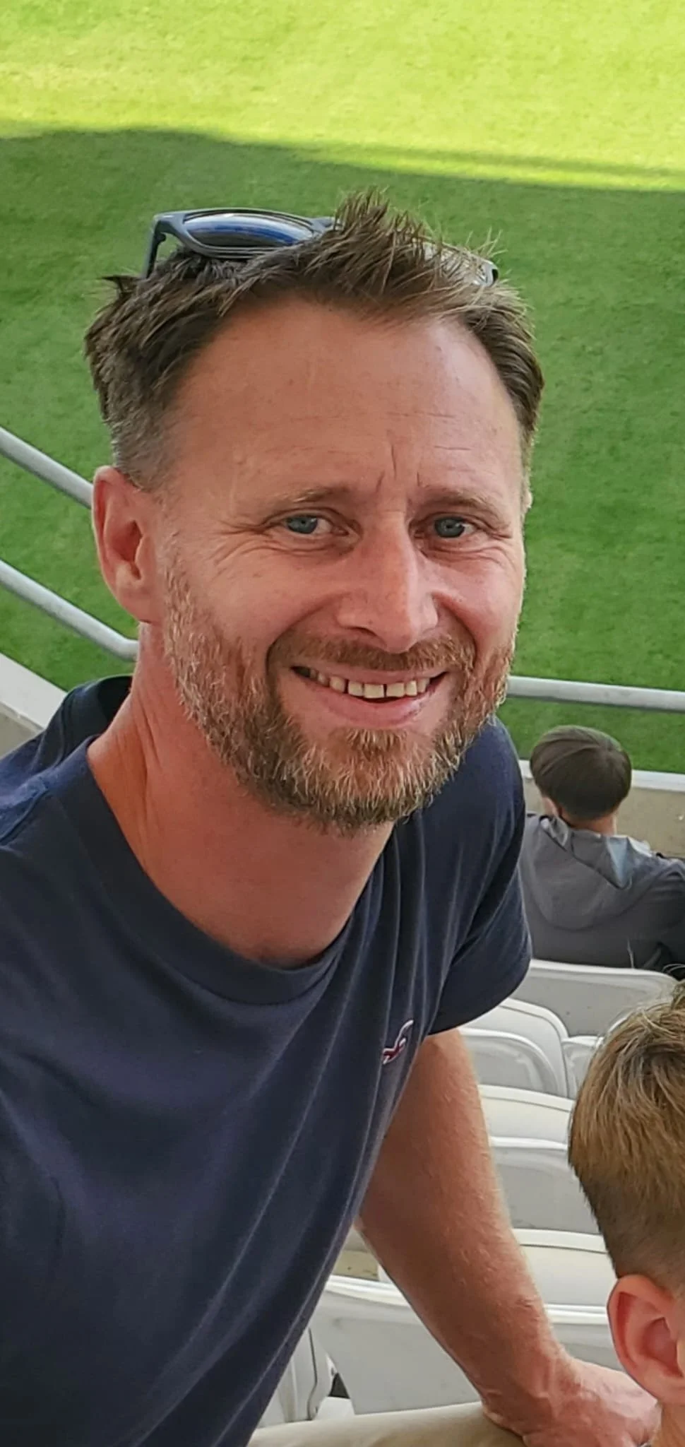 Smiling man with short brown hair, beard, and sunglasses on his head at a sports stadium, with green field and other spectators in the background.