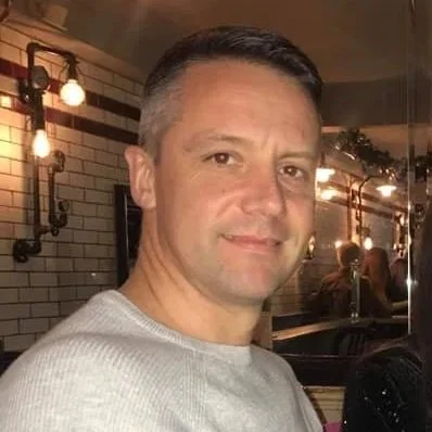 A man with short dark hair and light skin smiling in a restaurant or cafe setting, with warm lighting and brick walls in the background.