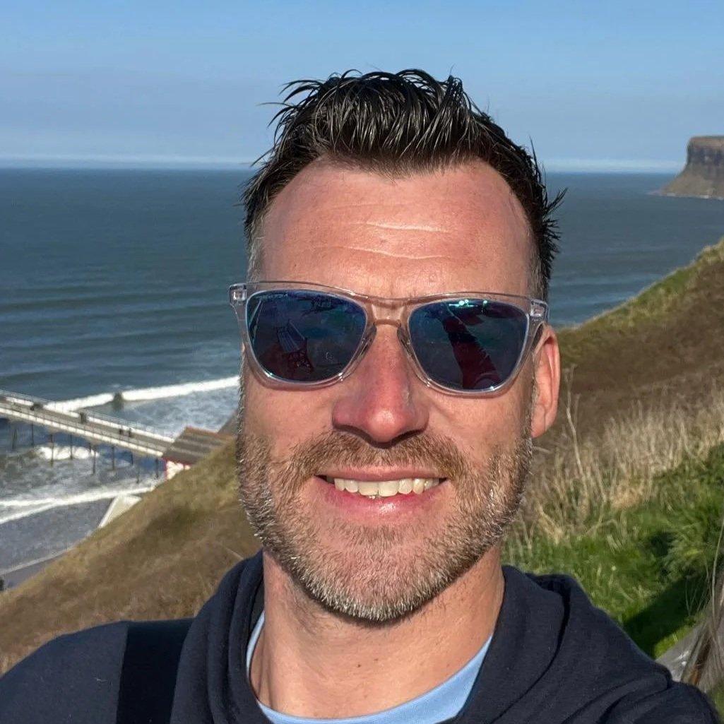 A smiling man with sunglasses taking a selfie on a hill overlooking the ocean with cliffs in the background.