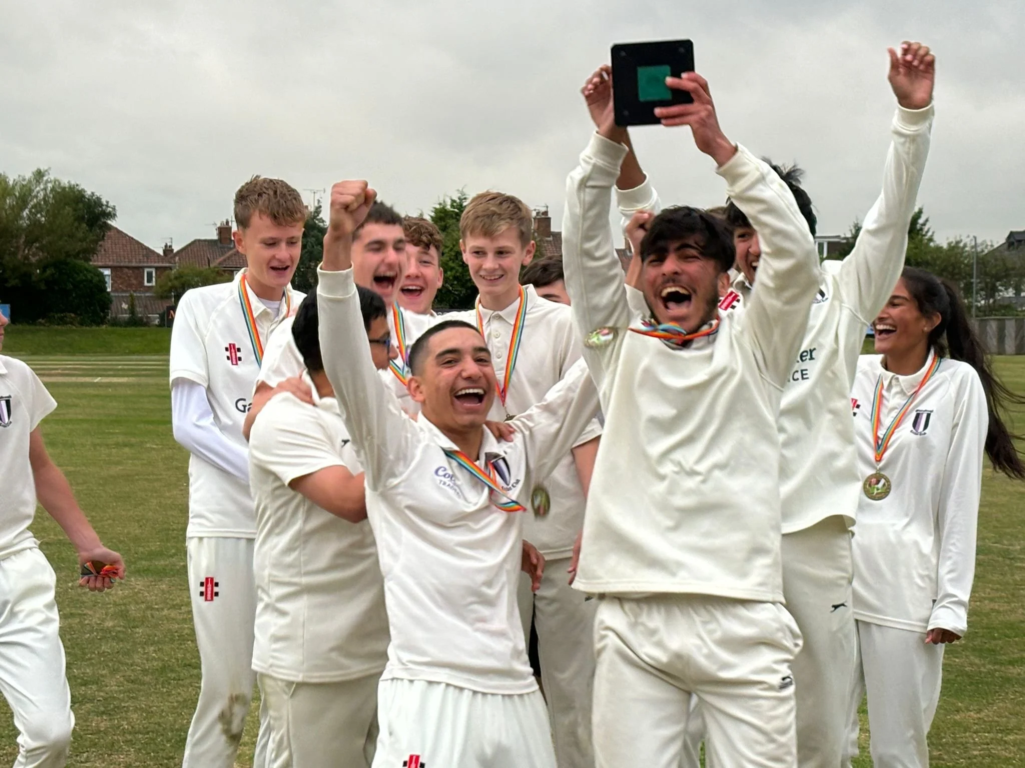 A group of young cricket players celebrating with medals around their necks on a grass field, taking a selfie. They are wearing white cricket uniforms and are smiling and cheering.
