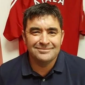 Close-up of a smiling man with dark hair wearing a dark collared shirt, with a red sports jersey hanging behind him.