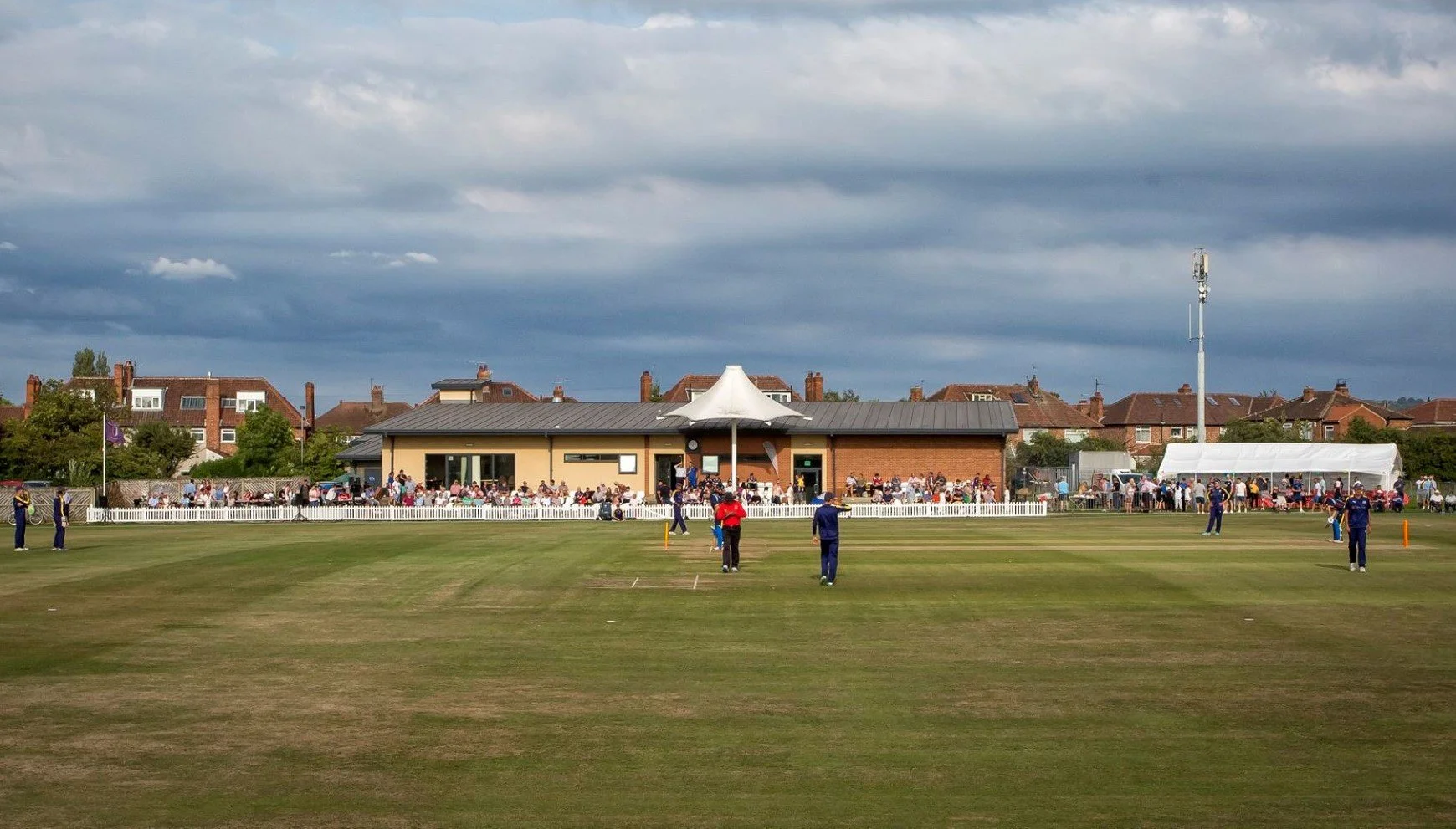 A cricket ground with players, spectators on benches and a building in the background under cloudy sky.