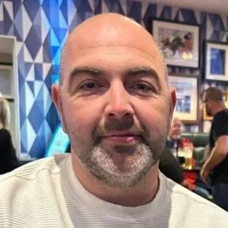 Close-up of a middle-aged man with a beard and bald head, wearing a light-colored shirt, in a restaurant with blue geometric wall art and framed pictures in the background.