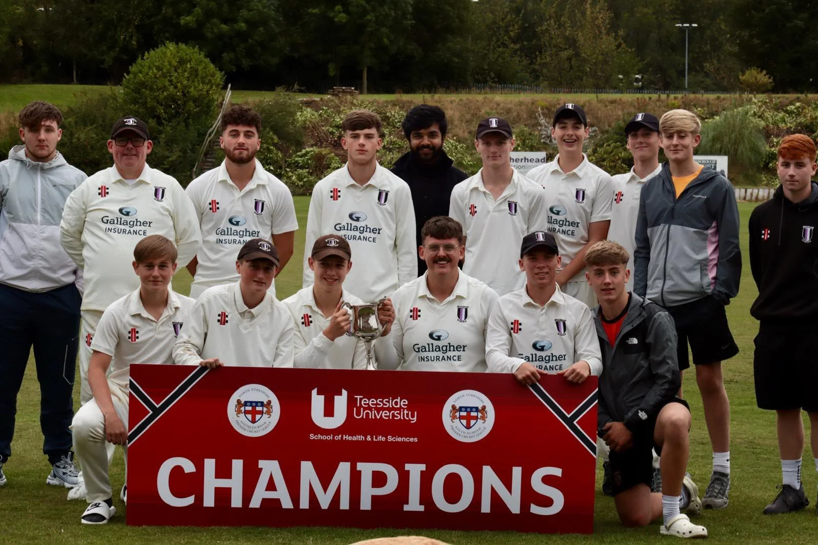 A group of young cricket players and coaches posing on a cricket field holding a red banner that reads "Champions" with the logos of Teesside University and Northern Yorkshire Cricket. The team is celebrating their victory, some are holding a silver trophy, and they are surrounded by greenery and trees.