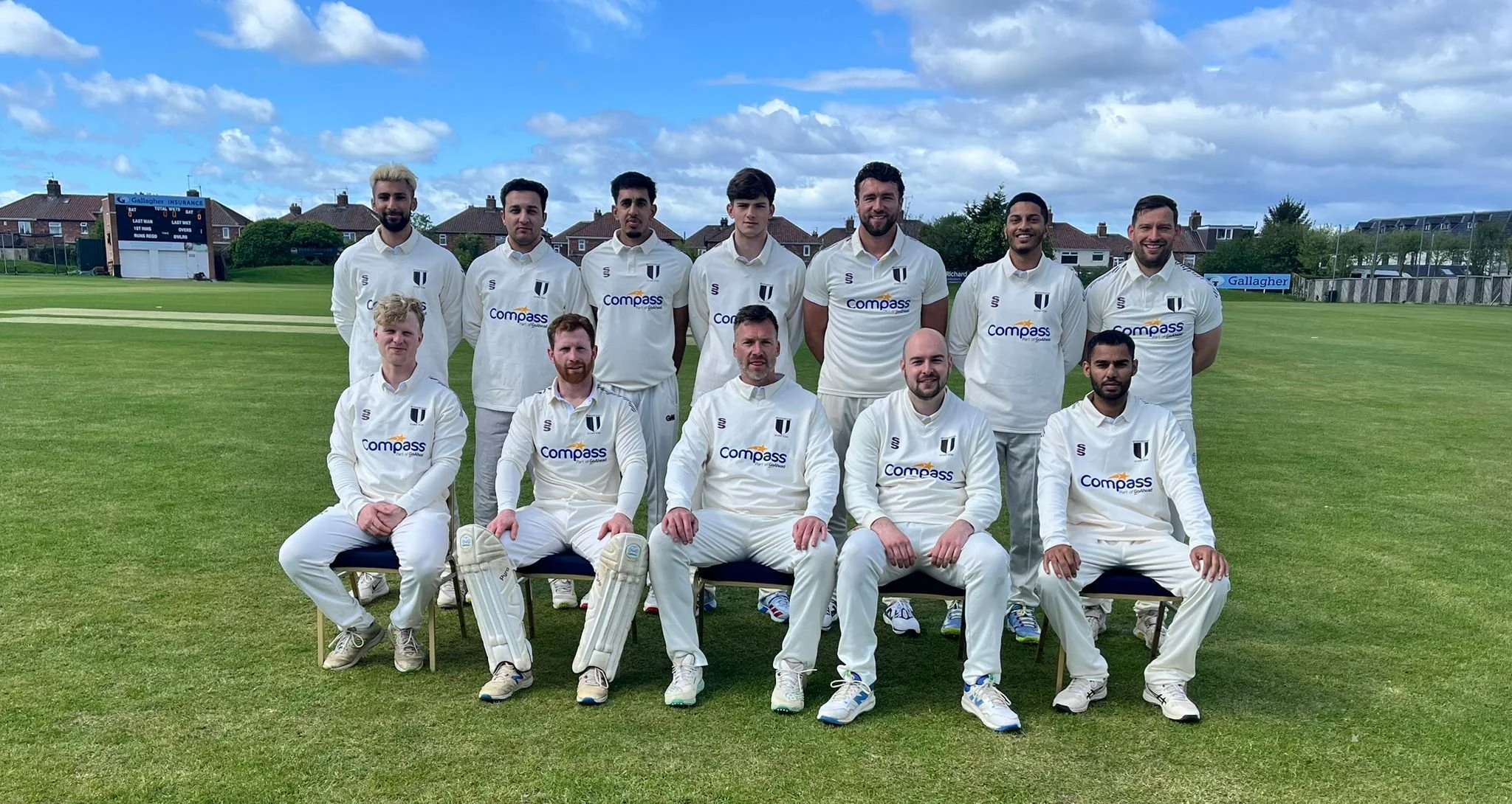 A cricket team posing on a grass field with a scoreboard and houses in the background. They are wearing white uniforms with the logo "Compass".