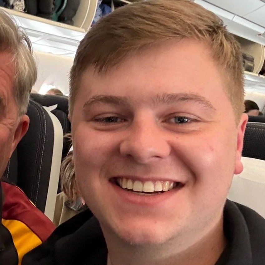 Smiling young man with short light brown hair in an airplane cabin, sitting next to a person wearing a maroon and yellow jacket.