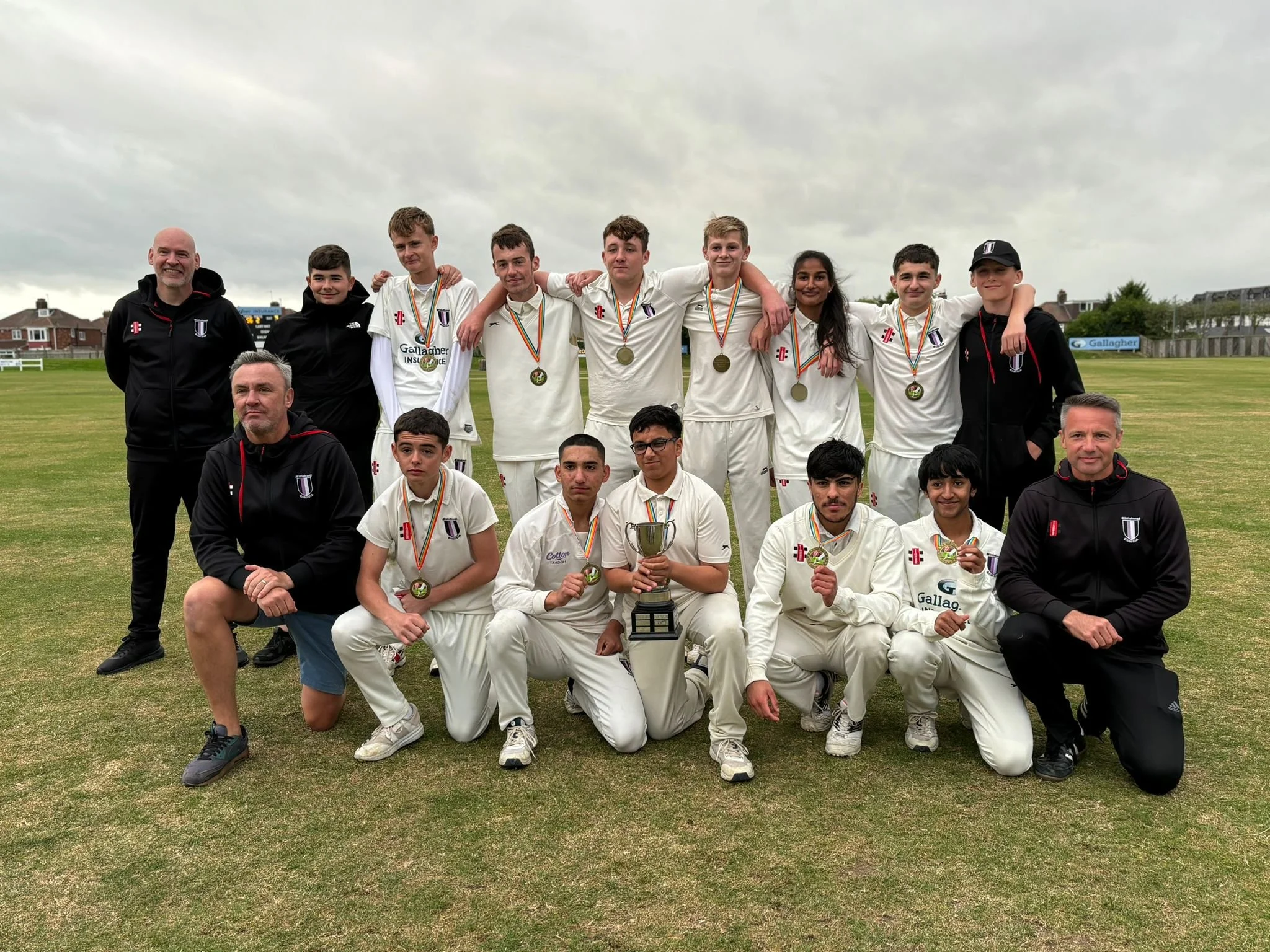 Cricket team with coaches celebrating victory, wearing medals, and holding a trophy on a cricket field.
