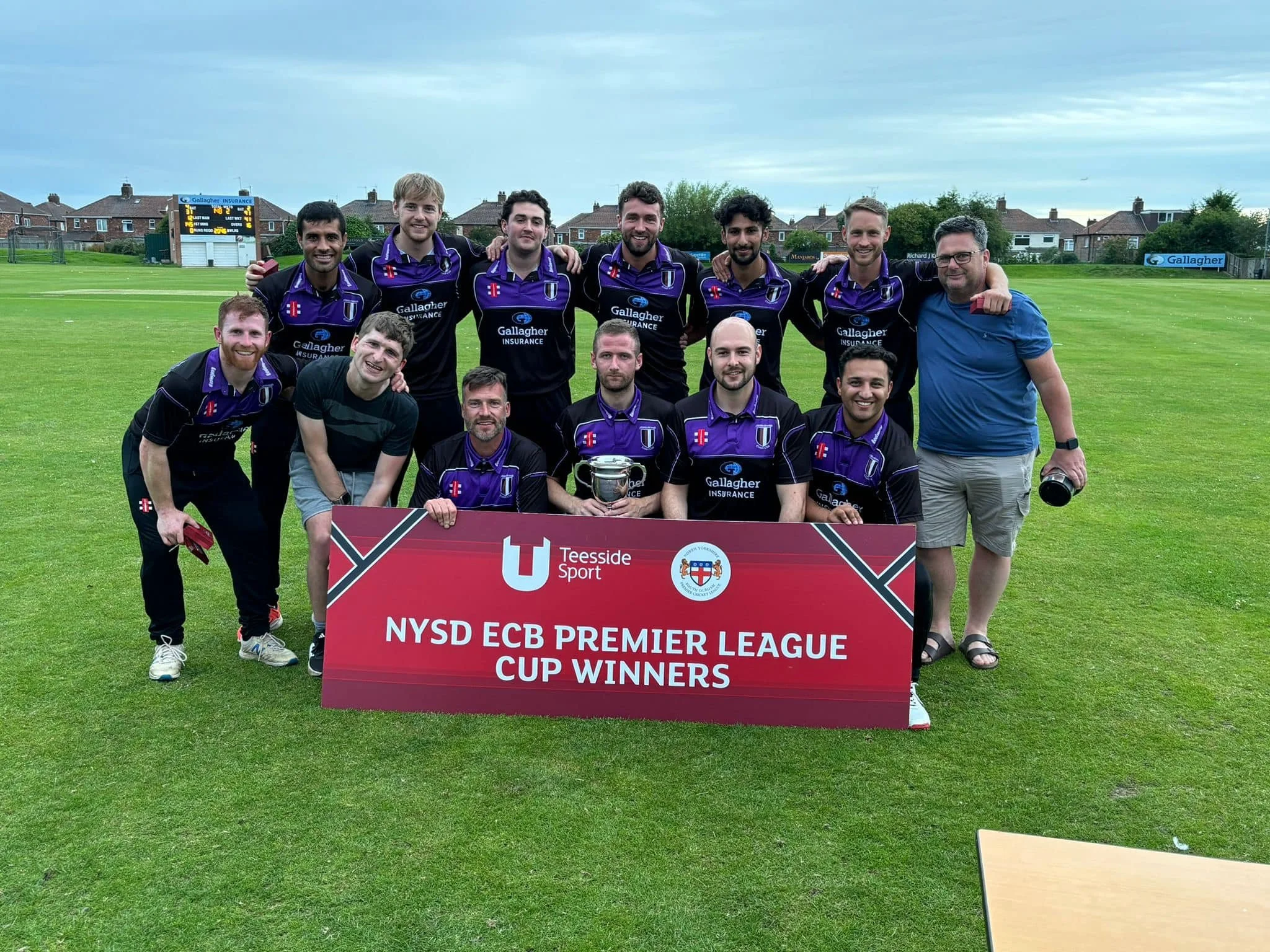 A group of eleven people, some in sports uniforms, pose on a grassy field holding a banner that reads 'NYS D ECB Premier League Cup Winners.' The team appears to be celebrating their victory in a sports event, with a scoreboard and residential houses visible in the background.