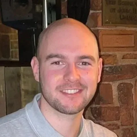 Close-up photo of a smiling young man with a shaved head and beard in front of a brick wall.