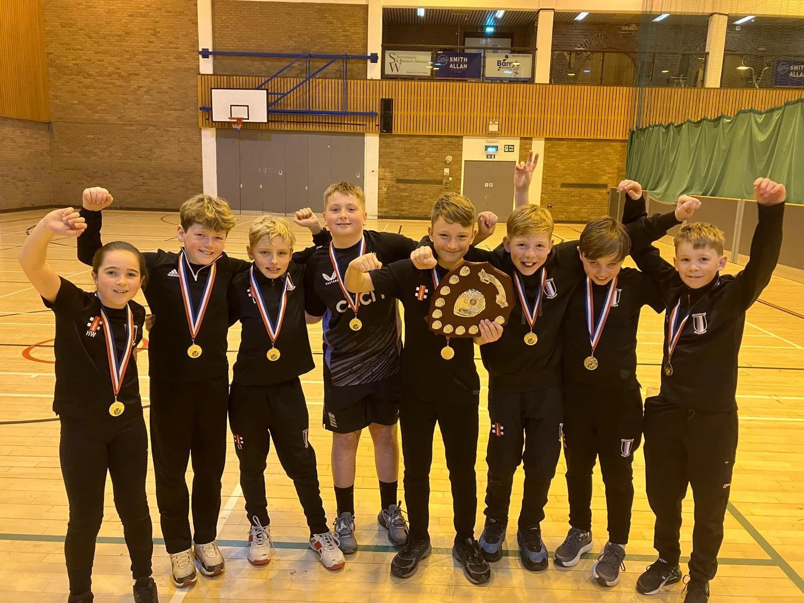 Young boys and girl in sports jerseys with medals around their necks, holding a trophy and celebrating in a gymnasium.