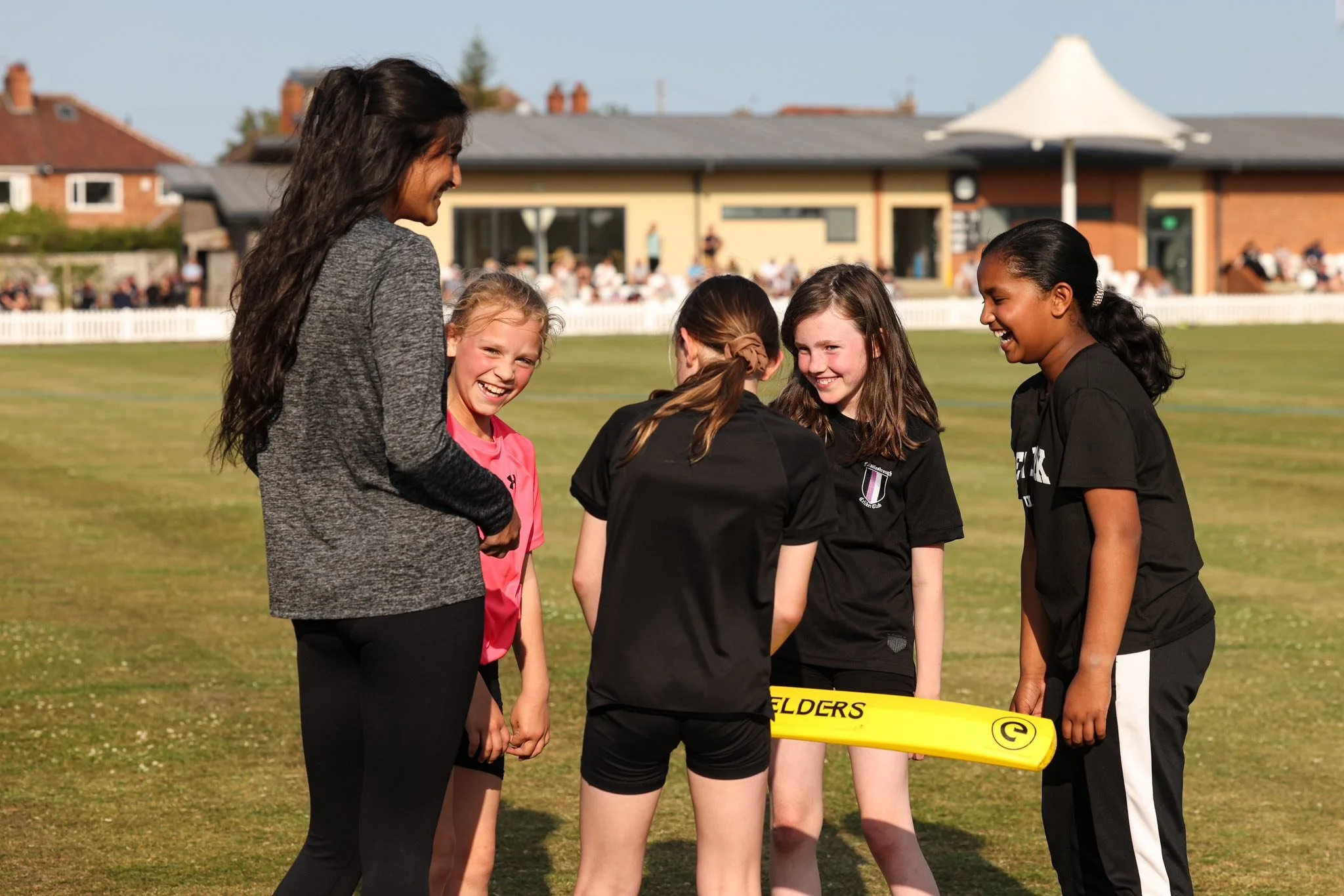 A woman and four young girls standing on a grassy field, laughing and smiling, holding a yellow relay baton, with a crowd and buildings in the background.