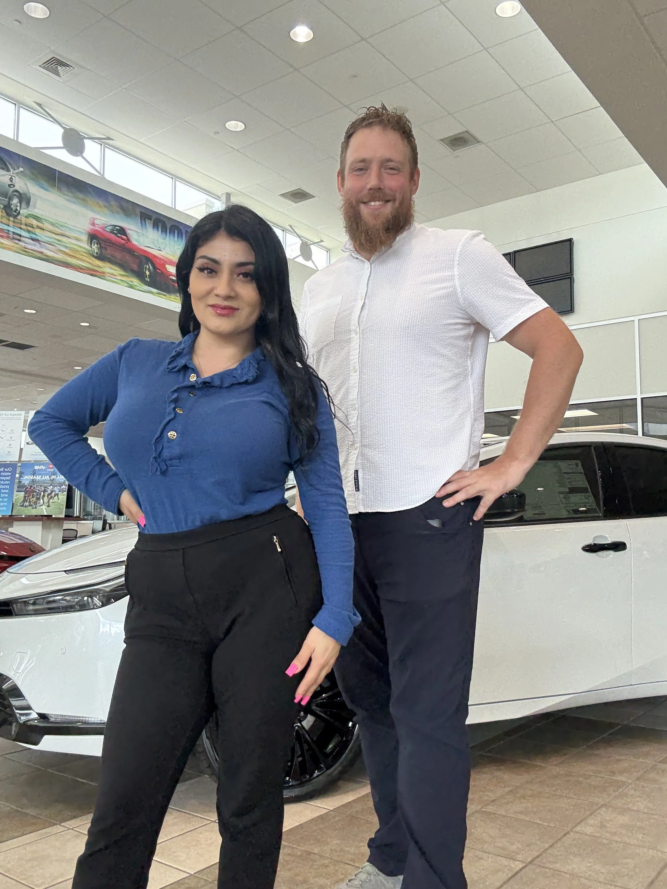 A woman and a man standing in a car dealership, with a white vehicle behind them. The woman has long dark hair and is wearing a blue top and black pants. The man has a beard, short curly hair, and is wearing a white button-up shirt and dark pants. Both are smiling and posing for the photo.