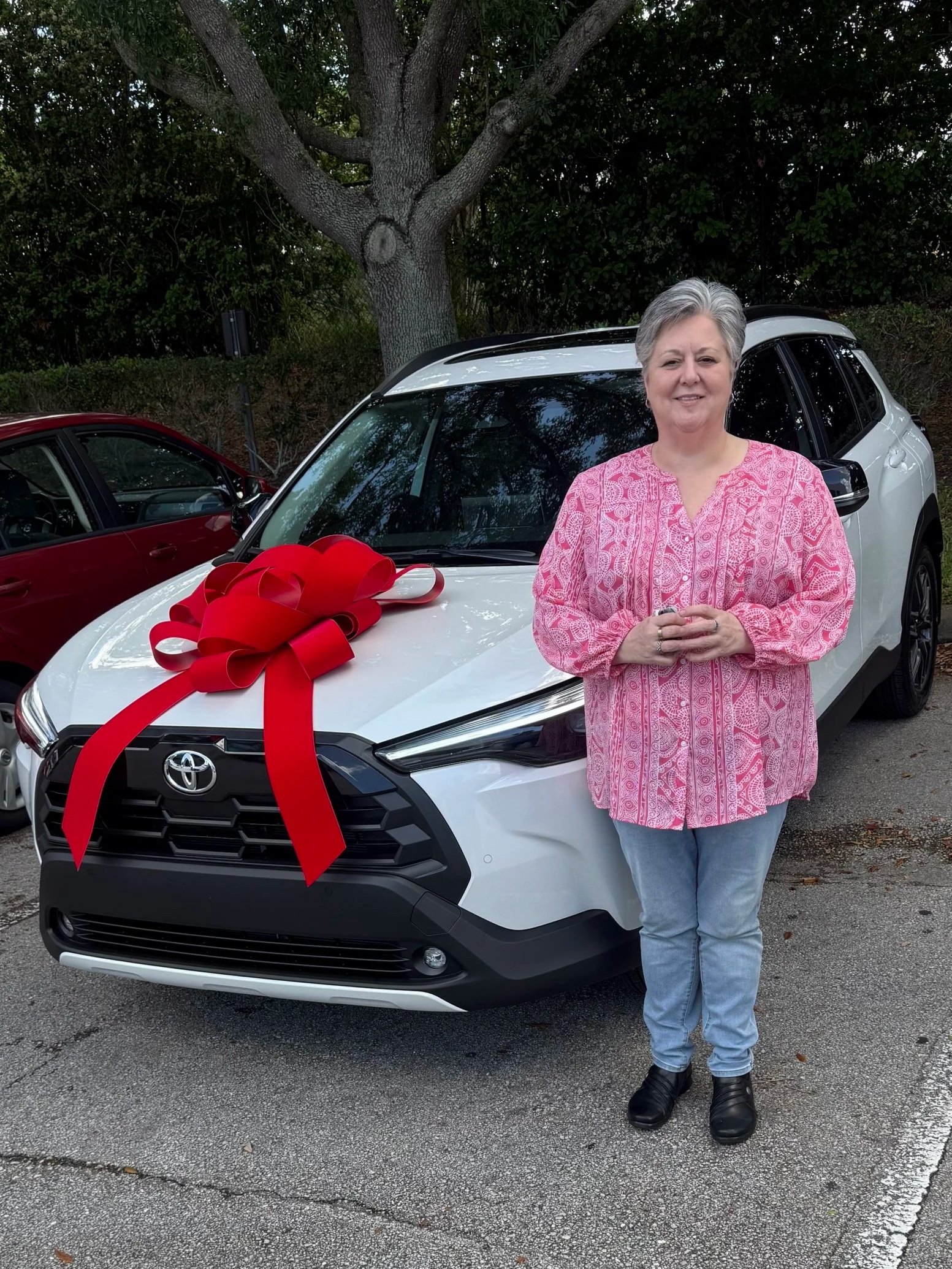 A woman standing in front of a white Toyota SUV with a large red bow on the hood, holding her hands together, outdoors with trees and parked cars in the background.