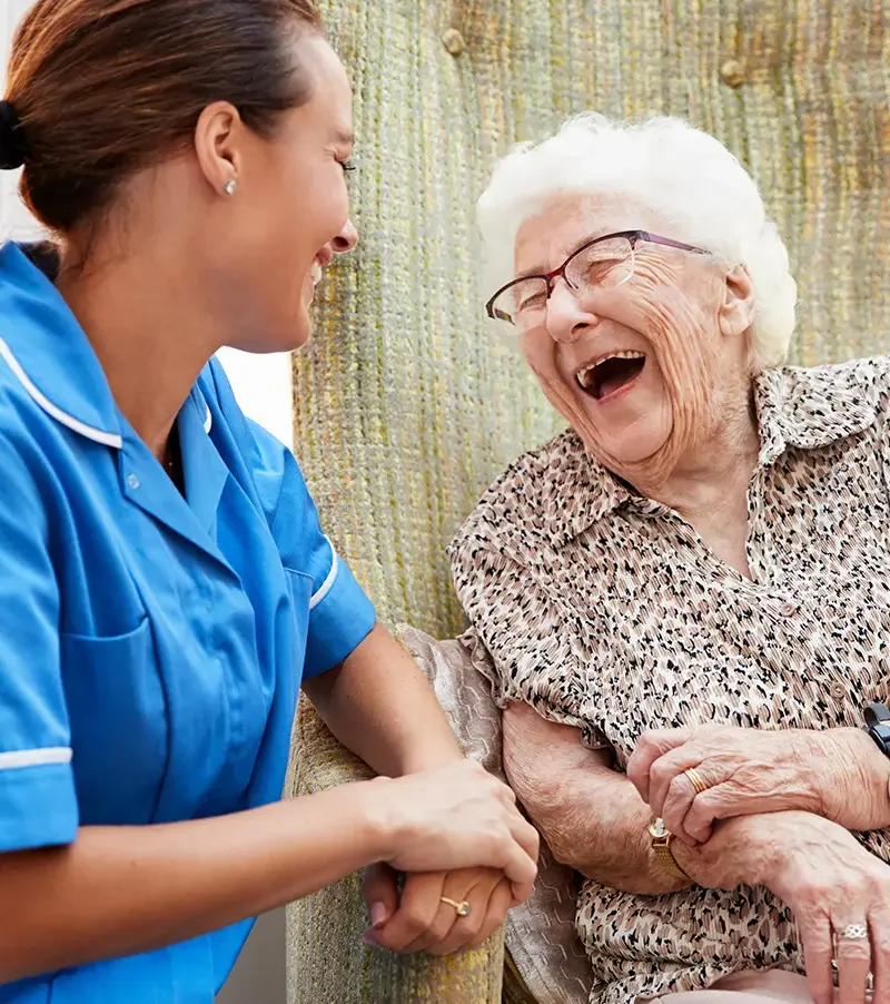 Personal Care - Home Care - Specialized Care - Disability Care - Post Hospital Care - Memory care - A young Rosebuds'  caregiver holding hands and smiling with an elderly woman with glasses and white hair, who is also smiling and laughing.