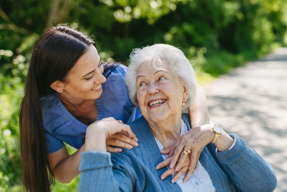 Personal Care - Home Care - Specialized Care - Disability Care - Post Hospital Care - Memory care - Dementia care - Companion  Care - A Rosebuds' caregiver caring for an elderly woman outdoors on a sunny day, smiling and holding her close.