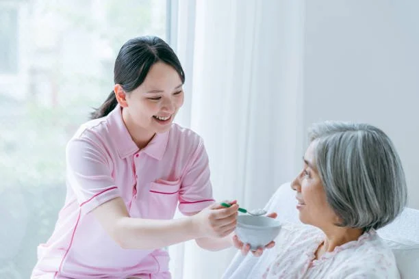 A young woman in a pink uniform feeding an elderly woman with gray hair in a hospital bed.