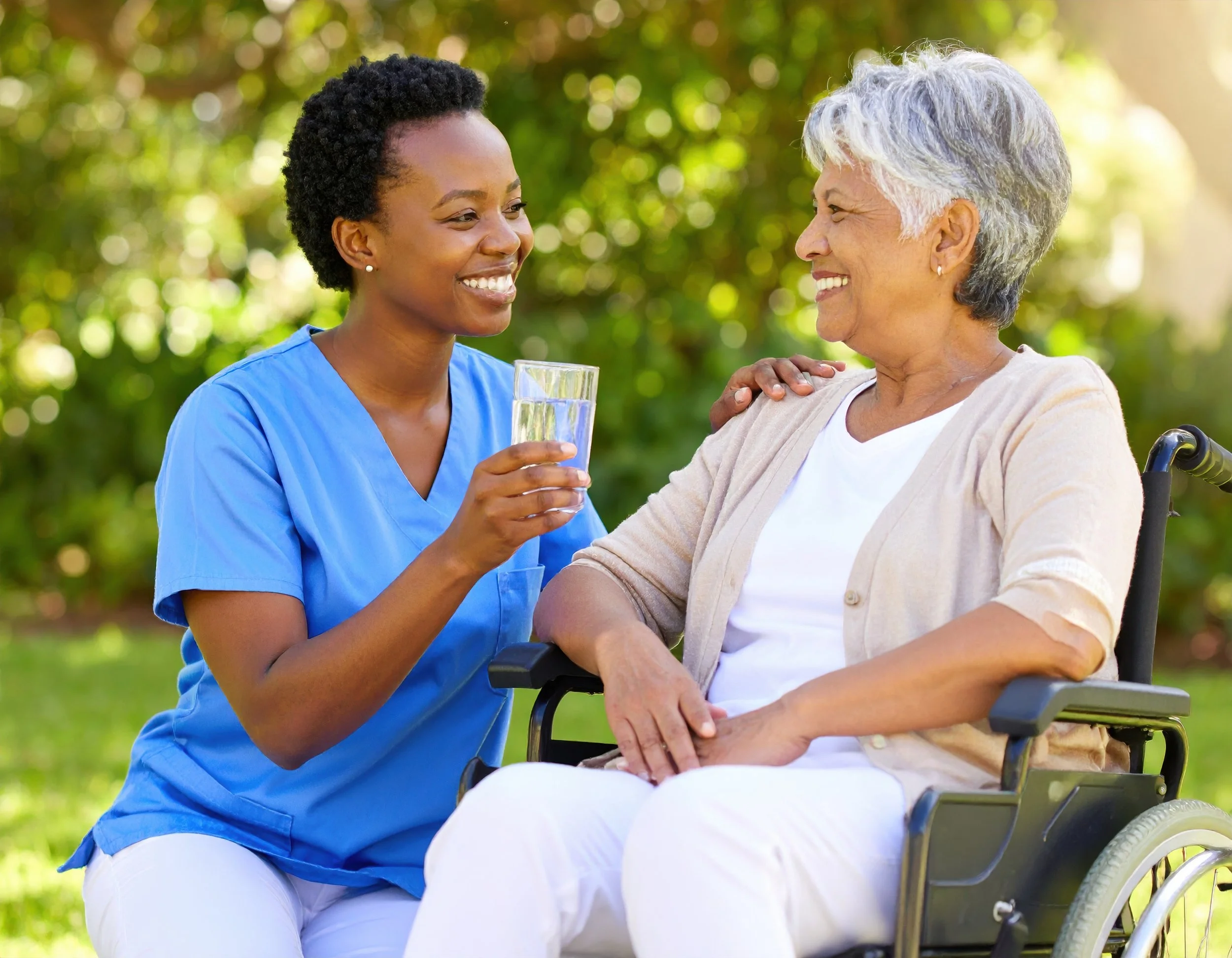 Personal Care - Home Care - Specialized Care - Disability care - A Rosebuds' caregiver smiling and holding a glass of water, talking to an elderly woman in a wheelchair who is smiling, outdoors with green trees in the background.