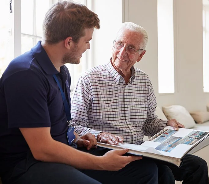 Personal Care - Home Care - Specialized Care - Disability Care - Post Hospital Care - Memory care - Dementia care - Companion  Care - A young Rosebuds' caregiver and an elderly man sit together, looking at a photo album and smiling.
