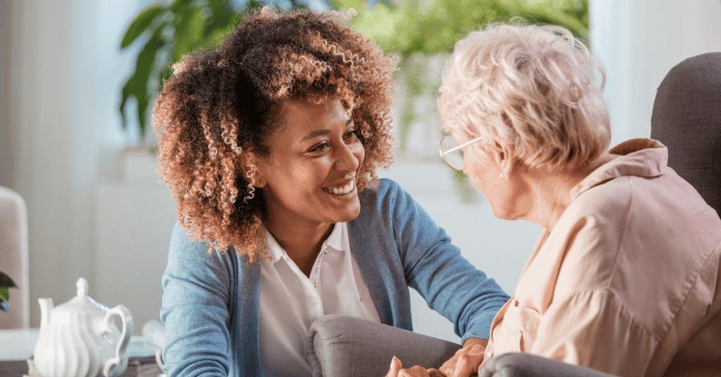 Personal Care - Home Care - Specialized Care - Disability Care - Post Hospital Care - Memory care - Dementia care - Companion  Care - 24/7 care - A Rosebuds' caregiver smiling and holding hands with an elderly woman in a cozy, well-lit room.