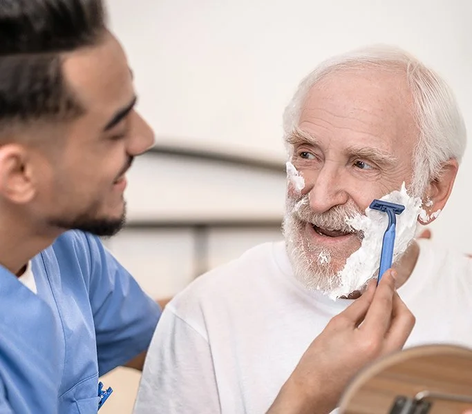 Personal Care - A Rosebuds male caregiver helping an elderly man shave using a razor, with shaving cream on his face.
