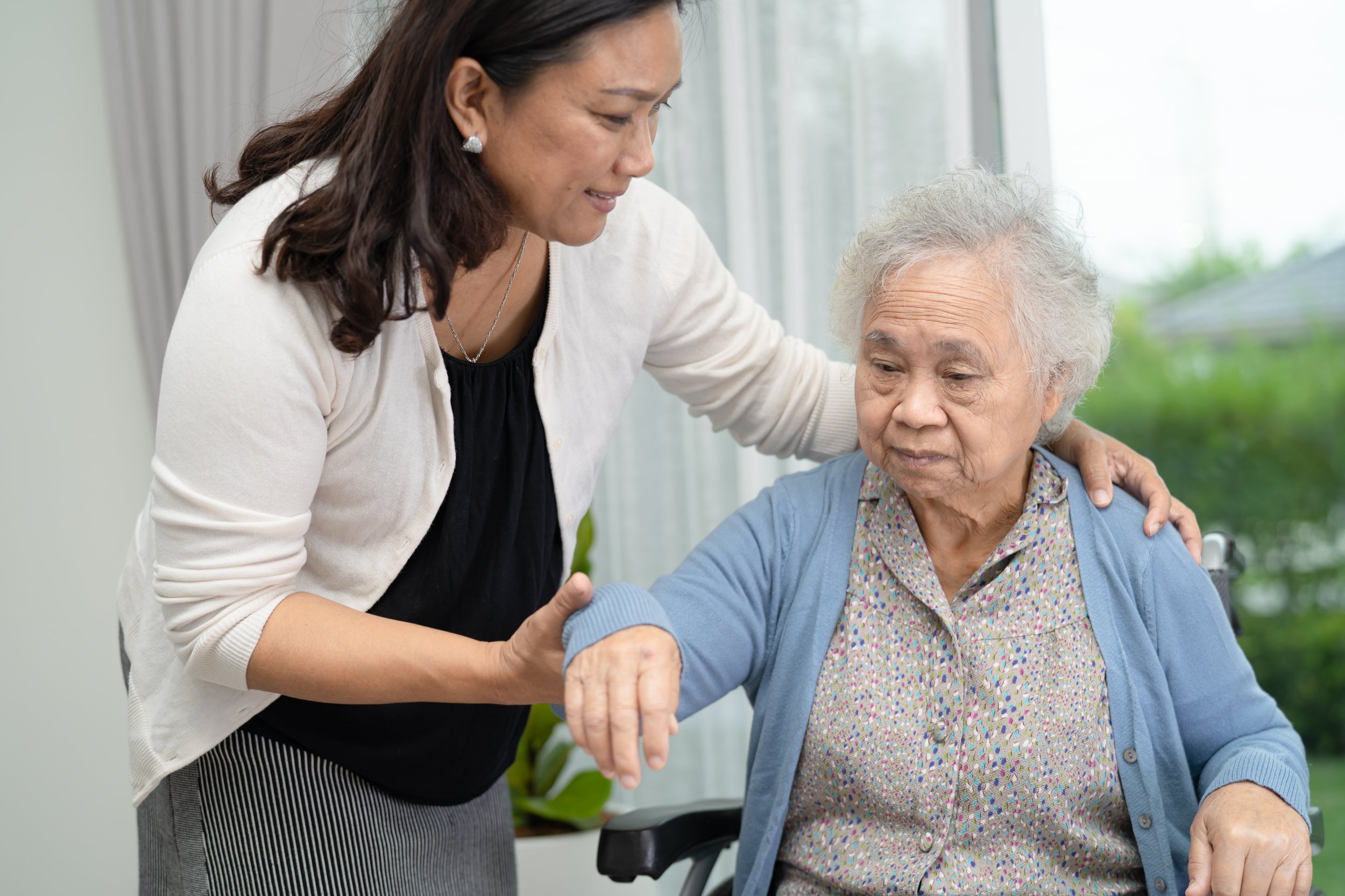 Personal Care - Home Care - Specialized Care - Disability Care - Post Hospital Care - Memory care - A Rosebuds'  caregiver assisting an elderly woman with her mobility, the woman sitting in a wheelchair and the caregiver gently holding her arm.