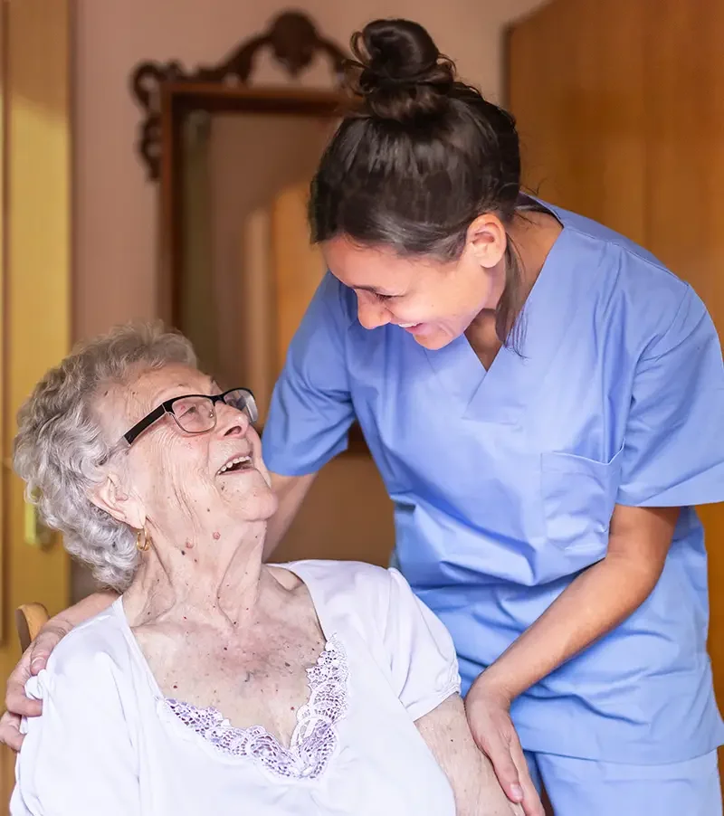 Personal Care - Home Care - Specialized Care - Disability Care - Post Hospital Care - Memory care - Companion care - A Rosebuds' caregiver comforting an elderly woman with glasses and gray curly hair, both smiling and sharing a joyful moment.