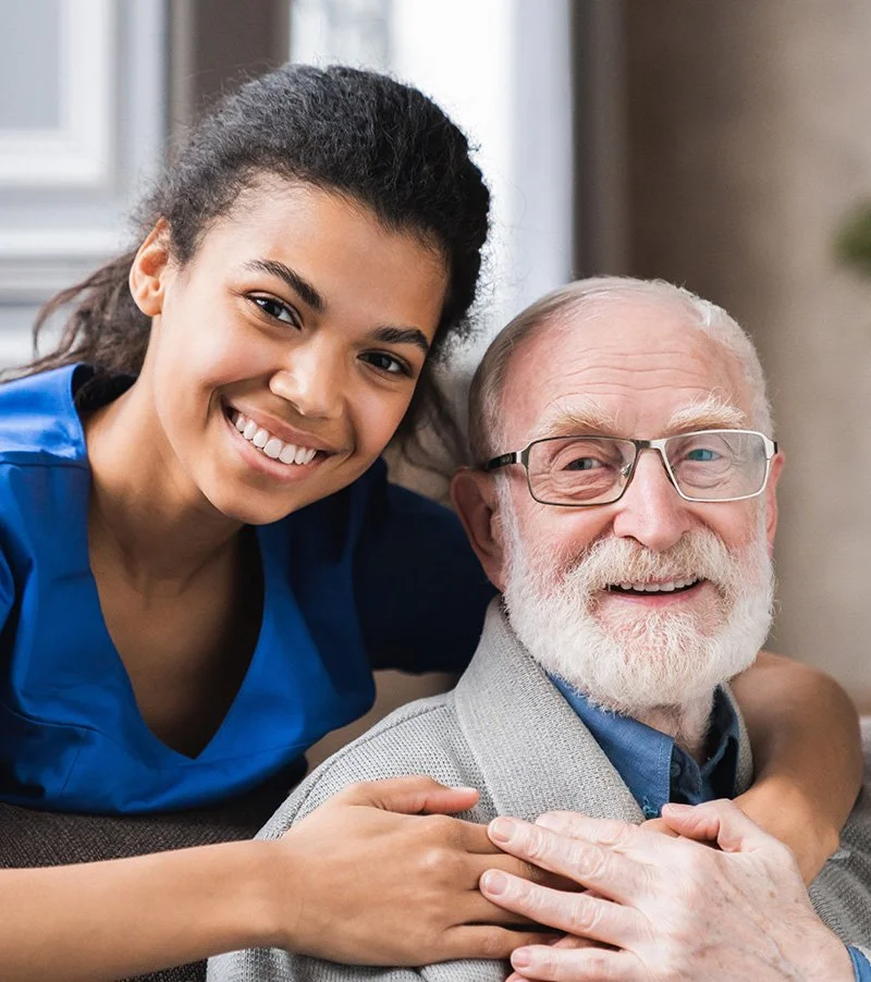 Personal Care - Home Care - Specialized Care - Disability Care - Post Hospital Care - Memory care - Dementia care - Companion  Care - 24/7 care - A Rosebuds' caregiver smiling and hugging an elderly man with a white beard and glasses.