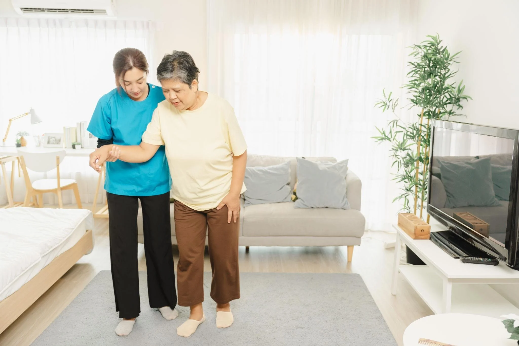 Personal Care - 24/7 Care - Live-In Care - Overnight Care - Specialized care - Companion care - Memory Care - Transitional Care - A Rosebuds' caregiver assisting an elderly woman with walking indoors, both wearing socks, in a well-lit living room.