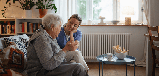 Personal Care - Home Care - Specialized Care - Disability Care - Post Hospital Care - Memory care - Dementia care - Companion  Care - 24/7/care - A Rosebuds' caregiver and an elderly woman sitting together in a cozy living room, smiling and talking.