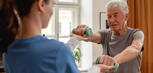 Personal Care - Home Care - Specialized Care - Disability Care - Post Hospital Care - Memory care - Dementia care - Companion  Care - 24/7 care-An elderly man lifting green dumbbells while assisted by a woman's trainer or therapist in a home setting.