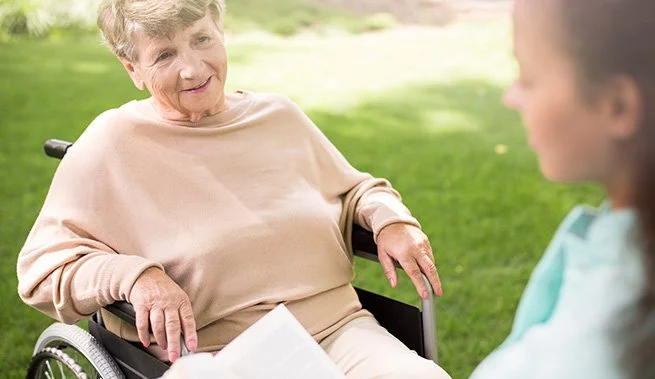 Personal Care - Home Care - Specialized Care - Disability Care - Post Hospital Care - Memory care - Dementia care - Companion Care - An elderly woman sitting in a wheelchair outdoors, smiling and engaging in conversation with a Rosebuds' caregiver.
