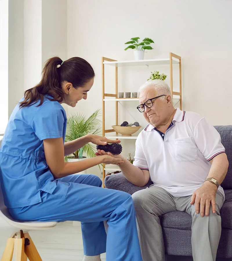 Personal Care - Home Care - Specialized Care - Disability Care - Post Hospital Care - Memory care - Dementia care - Companion  Care - A Rosebuds' caregiver shows a massage ball to an elderly man with glasses, sitting on a couch in a bright room.