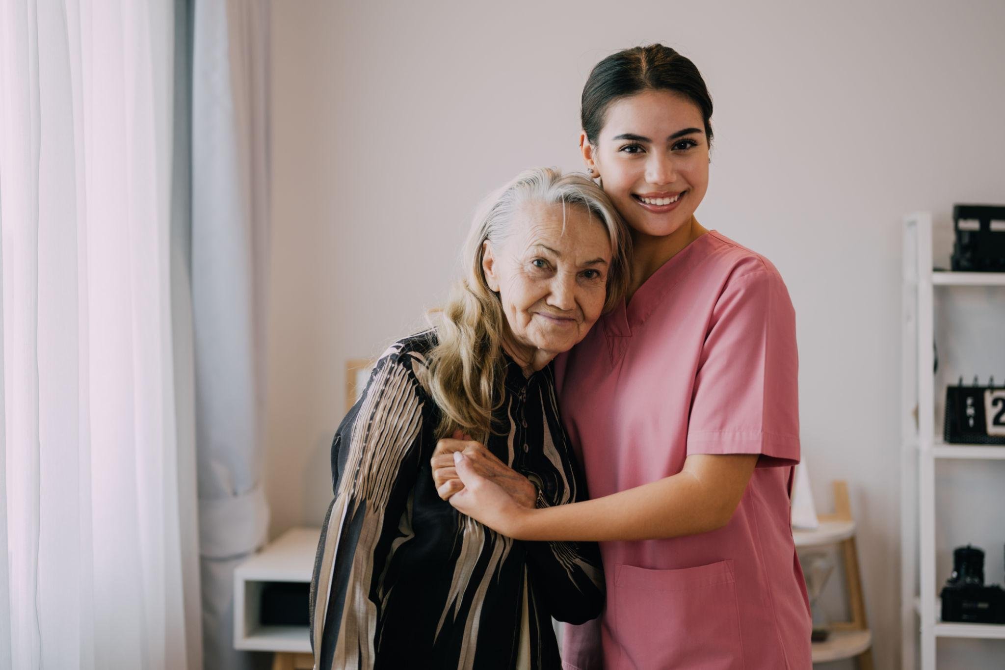 Compassionate Care - Personal Care - Companion Care - Memory care - Dementia care - Alzheimer's Care -  A young female Rosebuds' Caregiver in pink scrubs hugging an elderly woman with gray hair and a striped blouse inside a home.