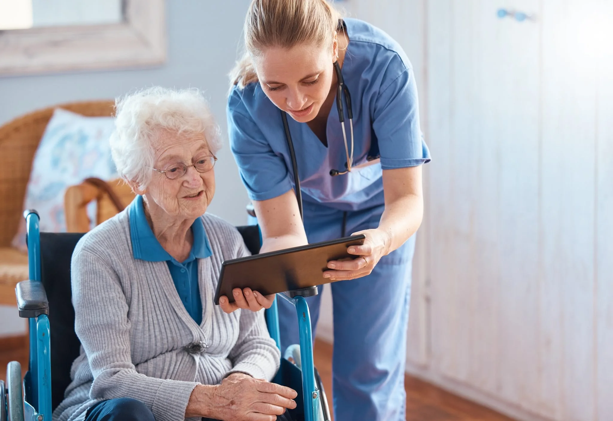 A healthcare worker in blue scrubs and a stethoscope shows an elderly woman in a wheelchair a tablet device inside a healthcare facility or nursing home.