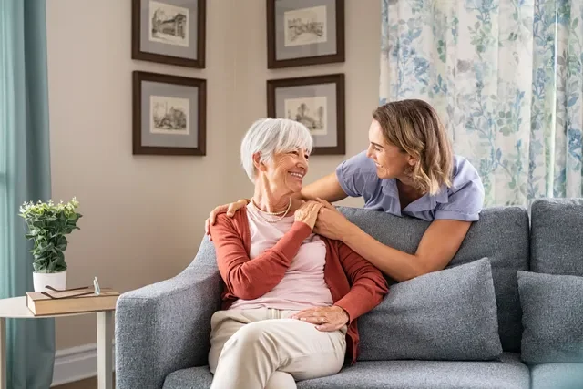 Companion Care - Memory care - Dementia care - Alzheimer's Care - Personal Care - A young Rosebuds' caregiver and an elderly woman sitting on a gray couch in a living room, smiling and engaging affectionately with each other.