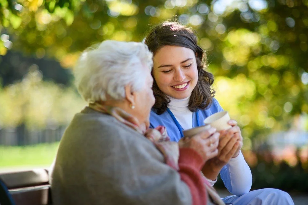 Personal Care - Home Care - Specialized Care - Disability Care - Post Hospital Care - Memory care - Dementia care - Companion  Care - A young Rosebuds' caregiver sharing a warm moment with an elderly woman outdoor, both holding bowls and smiling.