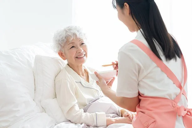 Specialized Care - Young female Rosebuds' caregiver smiling while feeding elderly woman in bed.