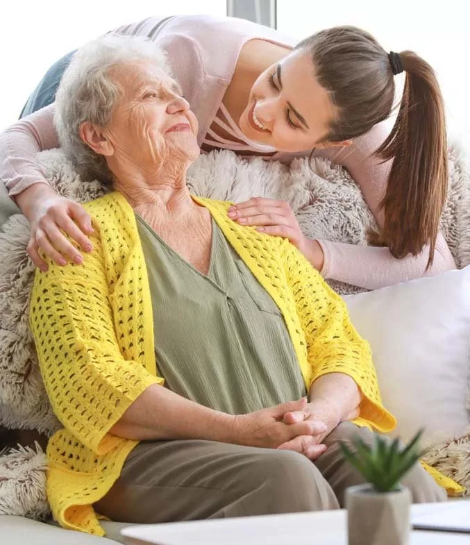 Personal Care - Home Care - Specialized Care - Disability Care - Post Hospital Care - Memory care - Dementia care - Companion  Care - Respite care - Live-In care - A young woman smiling and leaning over an elderly woman who is sitting on a sofa.