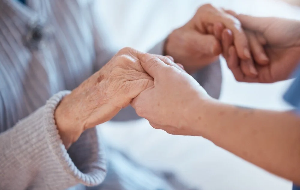 Personal Care - Home Care - Specialized Care - Disability Care - Post Hospital Care - Memory care - Dementia care - Companion Care - 24/7 care - Respite Care - Close-up of a Rosebuds' caregiver holding the hands of an elderly person.