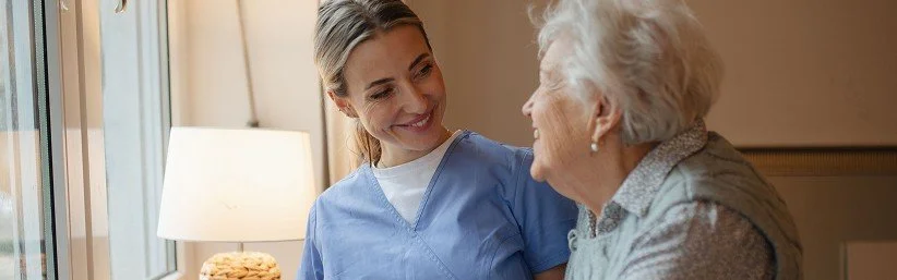 Personal Care - Home Care - Specialized Care - Disability Care - Post Hospital Care - Memory care - Dementia care - Companion  Care - 24/7 care - Rosebuds' caregiver assistant smiling and talking with an elderly woman in a care home.