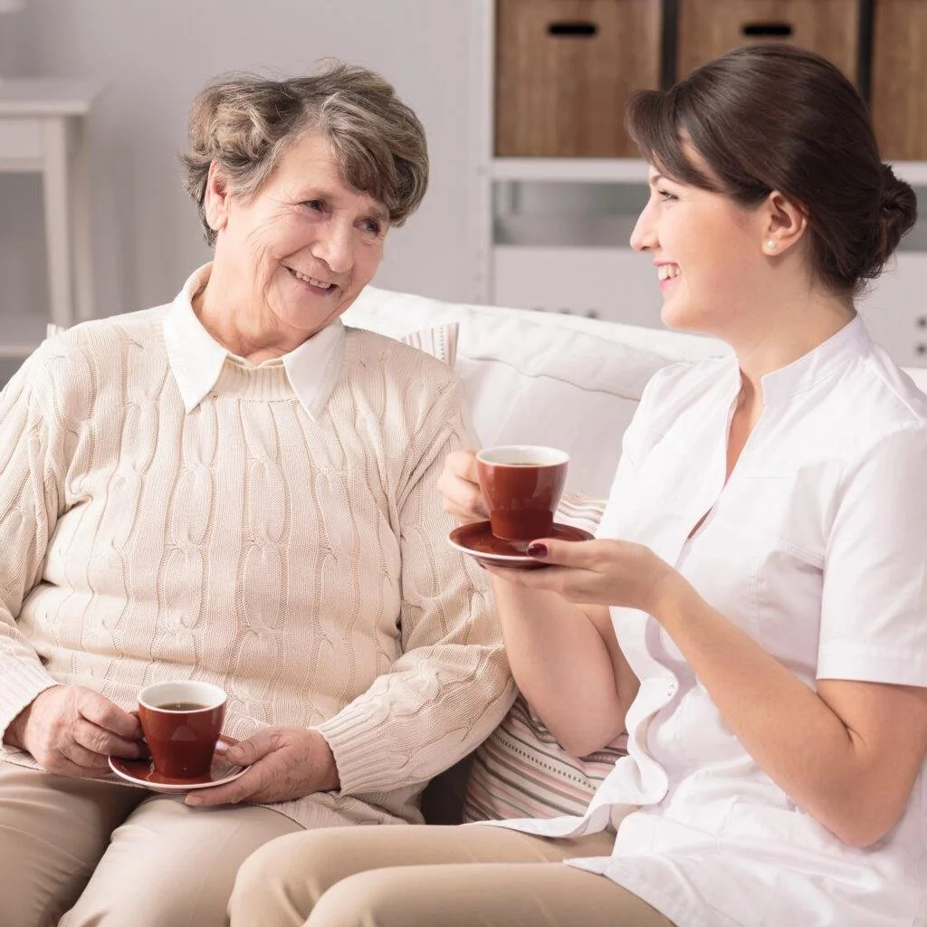 Personal Care - Home Care - Specialized Care - Disability Care - Post Hospital Care - Memory care - A Rosebuds' caregiver and an elderly woman share a cheerful moment together while sitting on a white sofa in a cozy room, each holding a cup of tea.