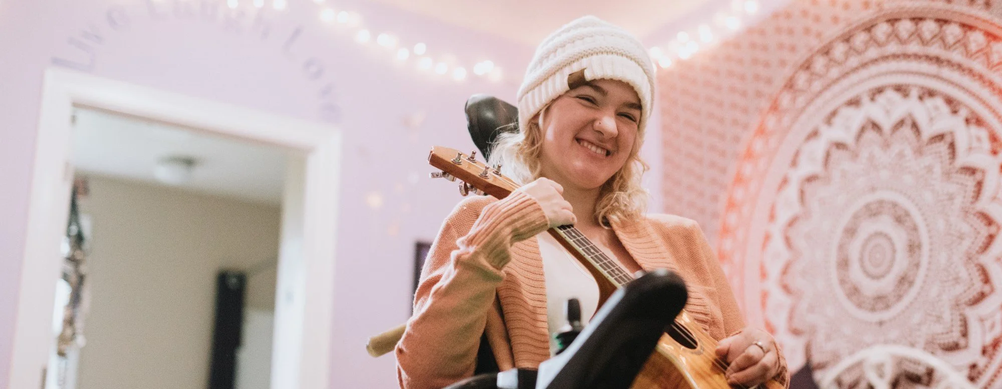 A woman smiling and playing an acoustic guitar in a cozy, decorated room with a pink and orange tapestry and string lights.